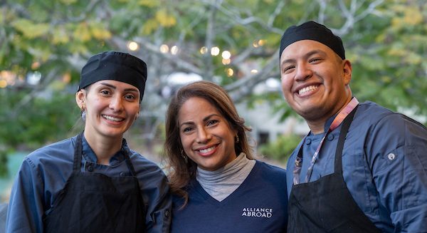 Three smiling hospitality professionals, including two chefs in black uniforms and a representative wearing an "Alliance Abroad" shirt, pose together outdoors in a warmly lit setting.