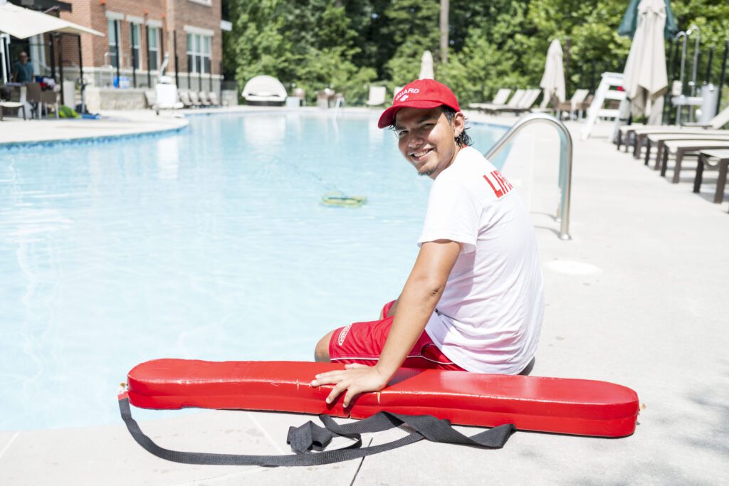 smiling lifeguard in a red cap and uniform sits by a pool, holding a rescue tube, during a sunny day at a summer work and travel program in the USA.