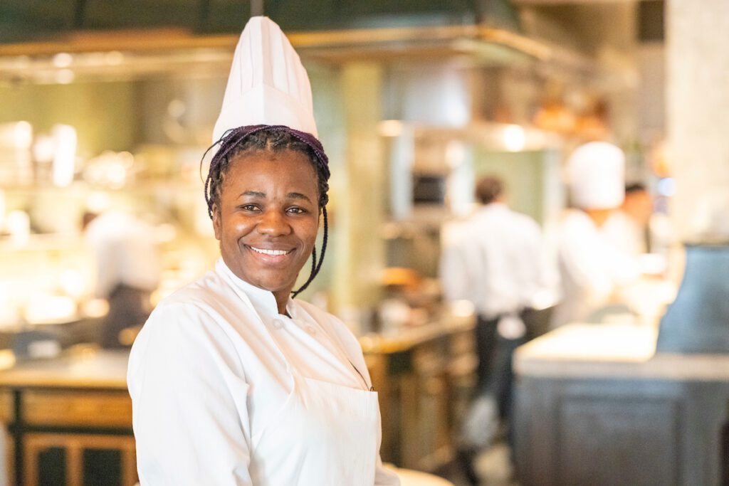 Smiling female chef wearing a white uniform and toque in a professional kitchen, with other chefs working in the background