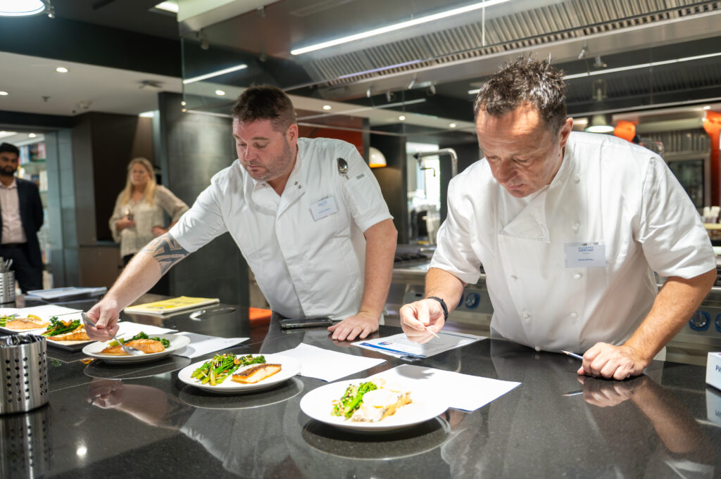 Two professional chefs in white uniforms carefully plating and evaluating dishes