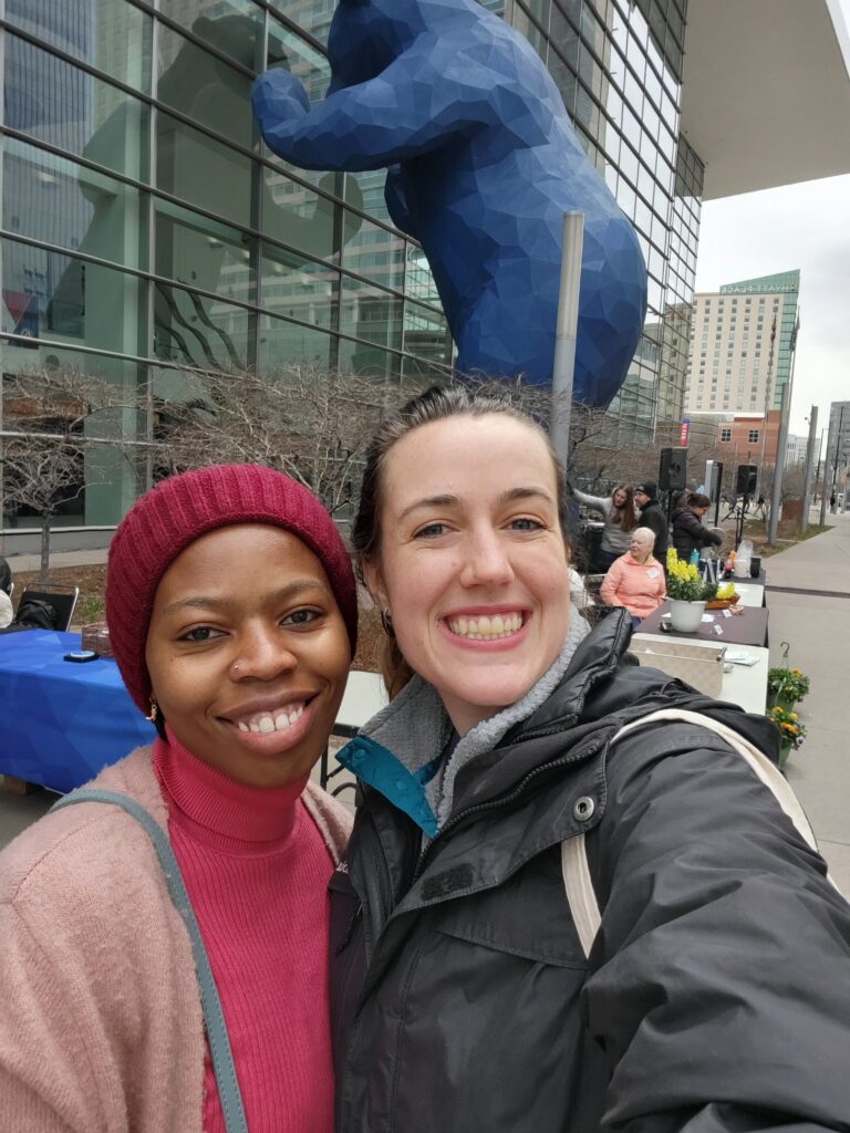 Two smiling women take a selfie in front of the iconic blue bear sculpture at the Colorado Convention Center, during an outdoor event showcasing culinary career opportunities abroad.