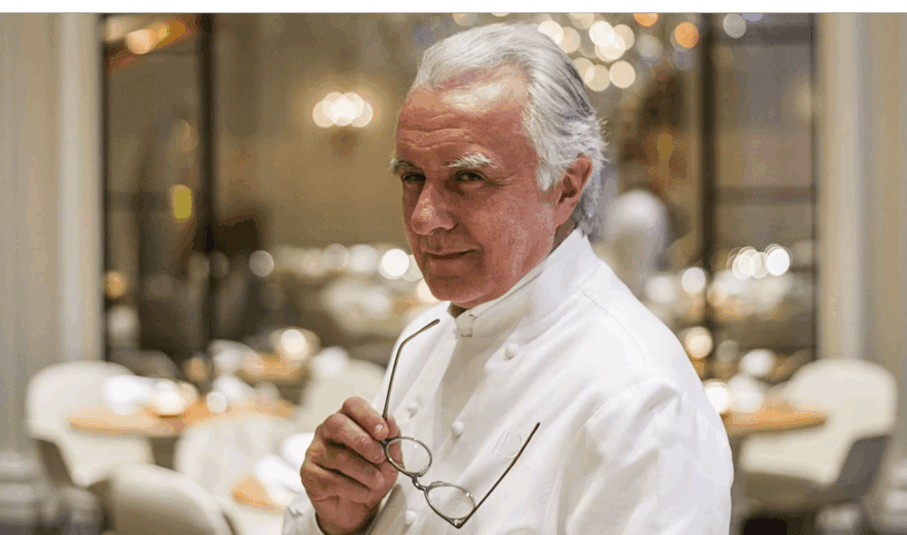 Alain Ducasse, renowned chef in a white uniform holding his glasses, standing confidently in a fine dining restaurant.