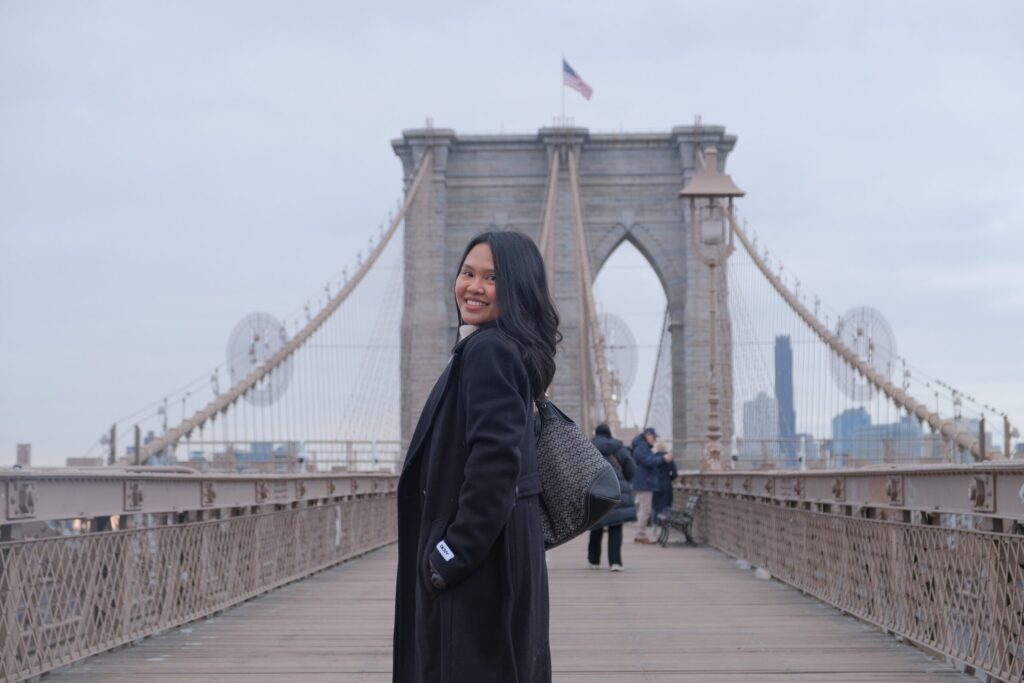 Participant Dindi Genil, smiling in front of the Brooklyn Bridge during her culinary journey abroad.