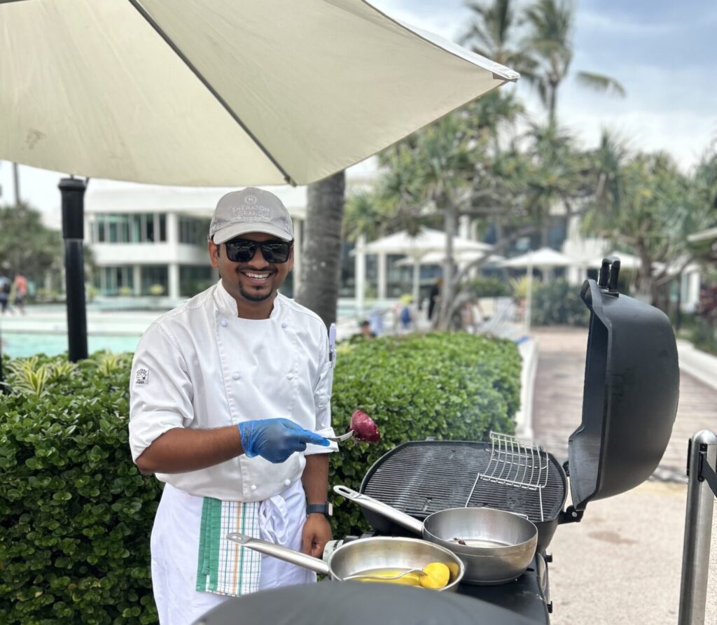 Participante en el programa Alianza en el Extranjero. Chef sonriendo mientras asa al aire libre en un complejo turístico bajo una gran sombrilla.