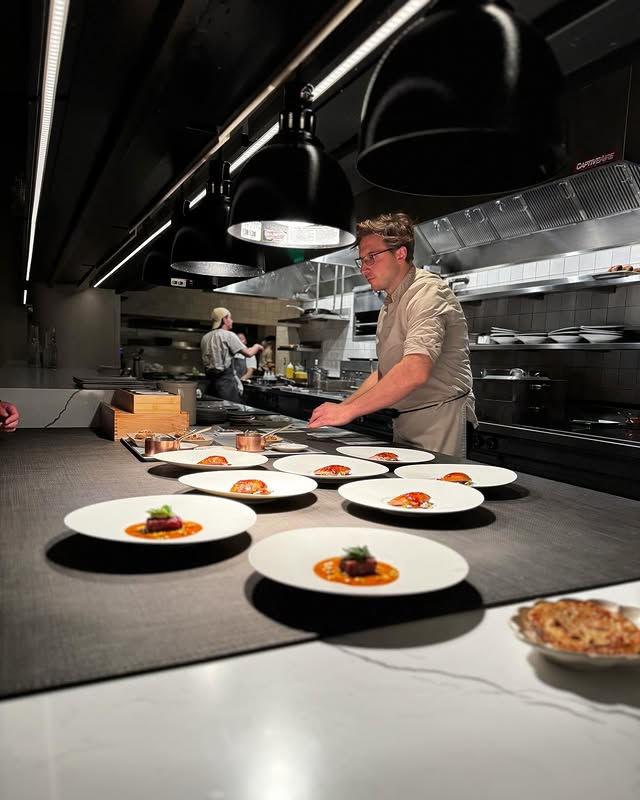 A chef plating multiple gourmet dishes in a modern open kitchen, showcasing fine dining techniques by some of the best chefs in the world in 2025.